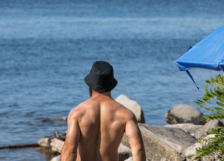 Back view of young handsome man standing at the beach and enjoying the sunshine. Portrait of muscular topless man with hat at the beach. travel photo, copy space for text, summer vacation concept.の写真素材