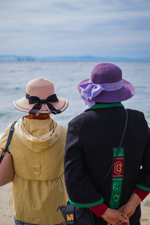 Two women in hats standing on the beach enjoying the view of the sea, back view. View of relaxed women by the seashore. Travel phot, copy space, selective focus.の写真素材