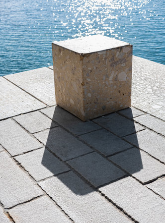 Concrete cube or cement brick with shadow. Abstract photo. Stone cube close-up with blue sea on the background. Massive ground cube. Nobody, street photoの写真素材
