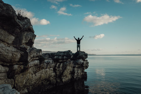 Back view of a man standing against the background of mountains with his arms outstretched. Young man stand on top of mountain top with hands raised in the air. Success and risk concept.の写真素材