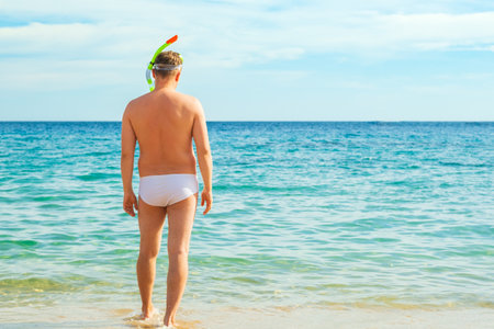 Rear view of an young man going snorkeling in the sea. Man goes snorkeling. Man standing on the beach going for a swim. Travel and holiday concept, copy spaceの写真素材