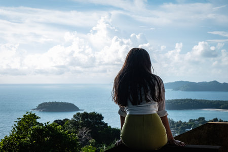 Back view of woman sitting and looking out towards blue ocean and sky. Young girl relaxing by the...