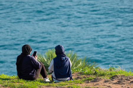 Two women in hijabs sitting on the grass. Back view of two female young friends outdoor by the sea.の写真素材