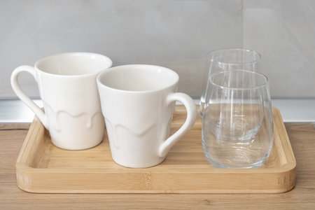 White cups for coffee or tea and drinking glasses. Two empty water glasses and two tea cups or mugs on a wooden tray in the kitchen, close up. Table wear, dishesの写真素材