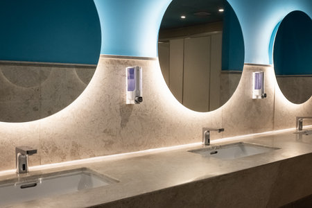 Modern toilet with a large round illuminated mirrors. Wide sink with taps. Interior of public toilet. Empty public restroom with washing hand sinks. Mirror with hand basin and faucet in clean toiletの写真素材