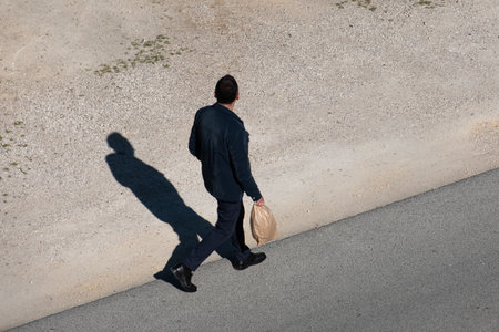 Man walking with the bag in his hands, rear view. Shadow of a man walking on the road in rural. Young man walking in the street on a sunny afternoonの写真素材