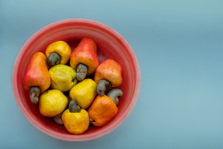 Ripe raw cashew nuts fruits harvested from the garden. Close-up view of colorful Cashew nut fruits in a bowl on light blue background. Nobody, selective focus. Farming, harvesting, agriculture conceptの写真素材