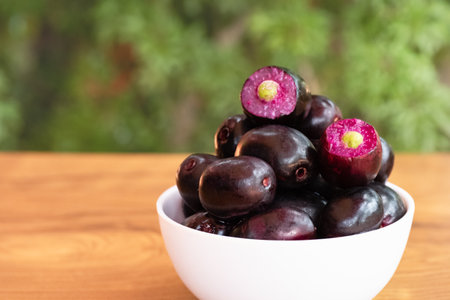 Jambolan plum with cut in half berries in a bowl. Jamun Black Plum Fruit in a Bowl on Wooden...
