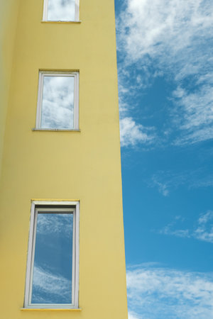 Reflection of a sky with clouds in the mirrored windows of a residential house. Modern building with windows and reflection of blue sky in them. Facade of bright colored building with blue skyの写真素材