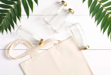 A reusable woven bag and glass bottles lie on a white background, near green leaves. Waste-free shopping set. Zero waste concept.の写真素材