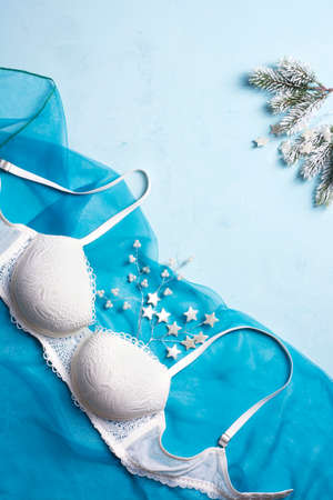 A beautiful lacy white bra lies on a blue fabric, near spruce branches and pearl jewelry. Concept for New Year, Christmas present. Flat lay, vertical orientation.の写真素材