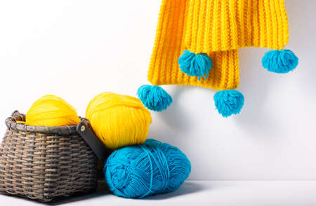 A wicker basket with yellow and blue yarn stands on a white background, with a yellow knitted scarf hanging in the background. Close-up, selective focus, horizontal orientation.の写真素材