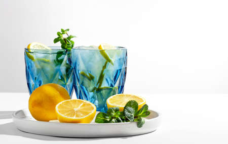 Two blue transparent glasses with cold lemonade stand on a white table, lemons, ice and mint lie next to it, selective focus on a lemon slice, close-up, copy spaceの写真素材