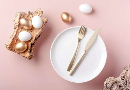 Easter white and gold eggs lie on the bark of a tree, next to a plate with cutlery, powdery background, horizontal orientationの写真素材