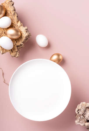 Easter eggs lie on the bark of a tree, next to an empty white plate on a powdery background, vertical orientation, copy spaceの写真素材