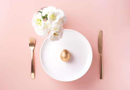 On a white plate lies a golden egg, next to white flowers, and golden cutlery on a powdery background, horizontal orientation, copy spaceの写真素材