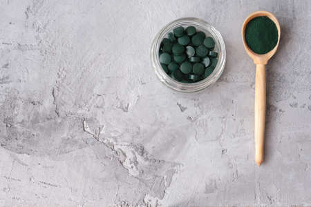 Green chlorella powder in wooden spoon and green chlorella tablets in small glass jar on gray concrete background. Chlorella is a single-celled green algae, it is used to make nutritional supplements and medicine. Top view.の写真素材