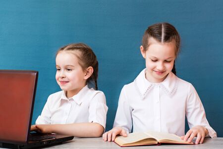 A girl works at a laptop and a girl reads a book at the table. Distance learning for children....