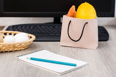 The concept of online grocery shopping without leaving home. Notepad for a shopping list on the background of a basket with eggs, a keyboard and a packet of food on the table.の写真素材