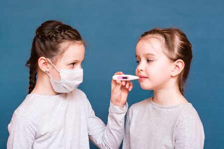 A girl in a medical mask measures the temperature of another with a thermometer in her mouth. Treating childrenの写真素材
