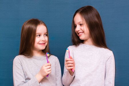 Two girls are smiling, looking at each other and holding toothbrushes in their hands on a blue background. The concept of oral hygiene in childrenの写真素材