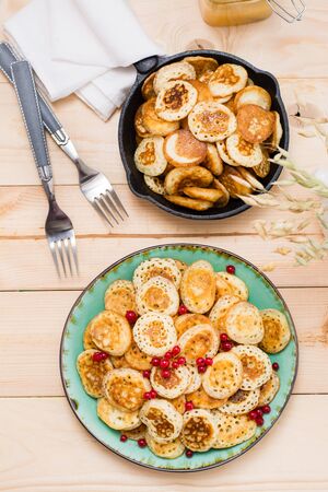 Trend breakfast. Dutch mini pancakes with red currants on a plate and a pan with them on a wooden table. Vertical. Top viewの写真素材