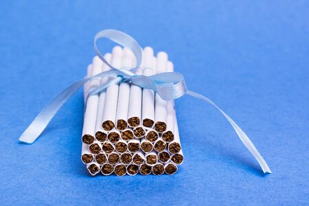 A stack of new thin cigarettes is tied with a blue ribbon with a bow lying on a blue background. The danger of smoking for women. Fatal addiction.の写真素材