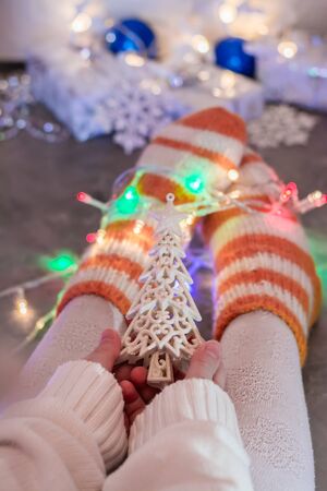 Christmas mood. A warmly dressed child holds in his hands a fir-tree ornament against the background of his legs in knitted socks, wrapped in a garland and wrapped gifts.の写真素材