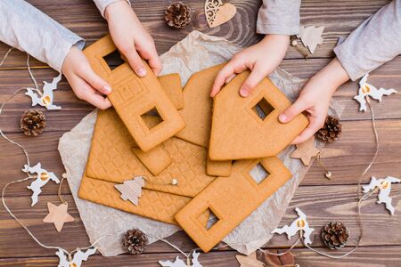 Parts of a disassembled Christmas gingerbread house in children's hands and Christmas decorations on a wooden background. Top view. Christmas preparationsの写真素材