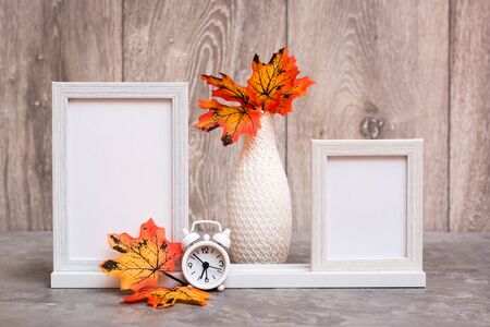 Ready Mock up. Two empty photo frames on a stand, a vase with orange maple leaves and a white alarm...