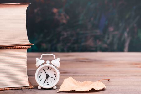 Back to school background with books, clock and fallen leaf on a wooden table over blackboardの写真素材