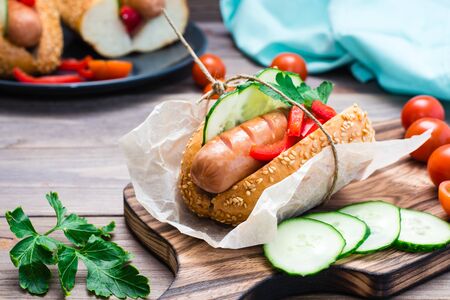 Ready-to-eat appetizing hot dog made from fried sausage, rolls and fresh vegetables, wrapped in parchment paper on a cutting board on a wooden tableの写真素材
