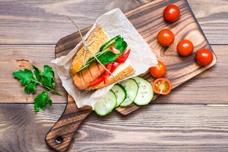 Appetizing hot dog made from fried sausage, rolls and fresh vegetables, wrapped in parchment paper on a cutting board on a wooden table. Top viewの写真素材