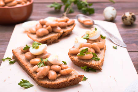 Bruschetta with boiled beans in tomato sauce, quail eggs and parsley on a cutting boardの写真素材