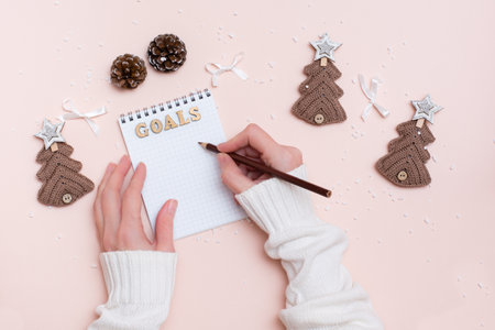 Year goals. Women's hands write with a pencil in a notebook and Christmas handmade on a pastel background. Knitted fir-trees, pine cones and bowsの写真素材