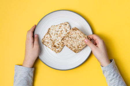 Healthy eating. Crispbread made from oats, wheat, flax and sesame seeds on a plate and hands on a yellow background. Top viewの写真素材