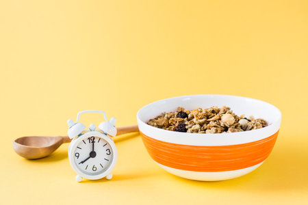 Healthy eating. Baked oats, nuts and raisins granola in bowl, wooden spoon and alarm clock on yellow backgroundの写真素材