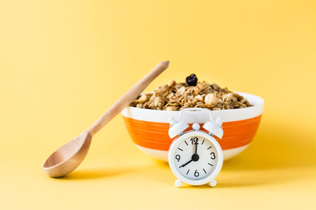 Healthy eating. Alarm clock in front of a baked granola made from oats, nuts and raisins in a bowl and a wooden spoon on a yellow backgroundの写真素材