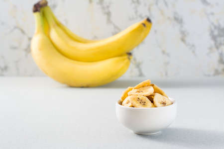 Baked banana chips in a white bowl and a bunch of bananas on the table. Fast food. Copy spaceの写真素材