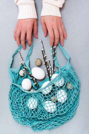 Happy Easter. Female hands hold a blue string bag with chicken and quail eggs and pussy willow branches on a gray backgroundの写真素材