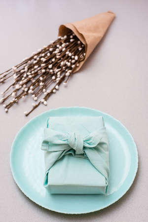 Eco-friendly gift - a box wrapped in cloth on a plate and a bouquet of pussy willow on a gray background. Minimalism. Vertical viewの写真素材