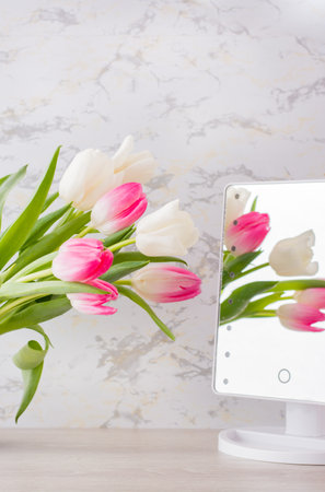 A bouquet of white and pink tulips with green leaves stands in a jug on the table and is reflected in the mirror. Selective focus. Vertical viewの写真素材