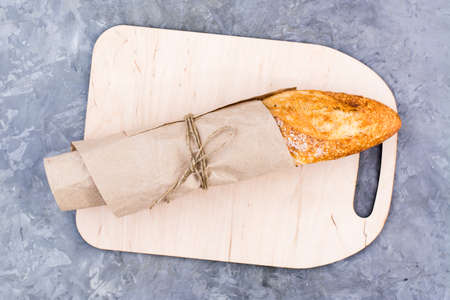Fresh baguette in wrapping paper on cutting board on table. Top view