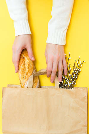 Food delivery concept. Female hands hold a paper bag with bread and a bouquet of pussy willow on a yellow background. Vertical viewの写真素材