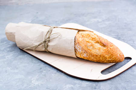 Fresh baguette in wrapping paper on cutting board on tableの写真素材