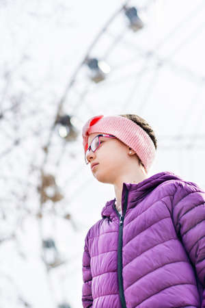Serious girl in glasses in early spring in a city amusement park against the background of a ferris wheelの写真素材