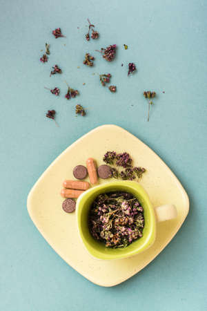 A cup with dried oregano and herbal medicinal capsules and pills on a saucer on a green background....