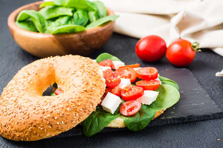 Appetizing bagels stuffed with tomatoes, feta and spinach leaves on a slate board on a black...