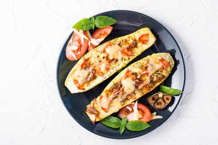 Ready-to-eat baked zucchini halves filled with cheese and tomato and basil leaves on a black plate on a white table. Vegetable menu, healthy food. Top viewの写真素材