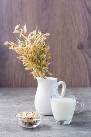 Oat milk in a cup, oatmeal and ears of corn in a jug on the table. Alternative to cow's milk. Healthy dietary non-allergenic food. Vertical viewの写真素材